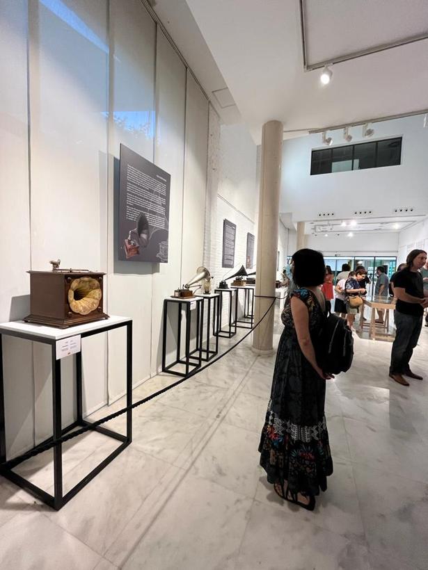 Sala de la exposición Sono-Art: gramófonos y fonógrafos sobre peanas negras a lo largo de una pared blanca, con público el día de la inauguración